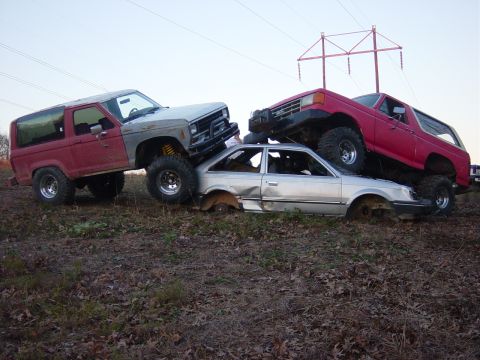 1983 Ford Bronco - Two off-road vehicles are positioned precariously on top of a severely damaged car.
