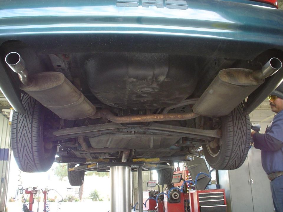 Underside view of a car's rear axle, dual exhaust pipes, and fuel tank. A mechanic is visible on the right