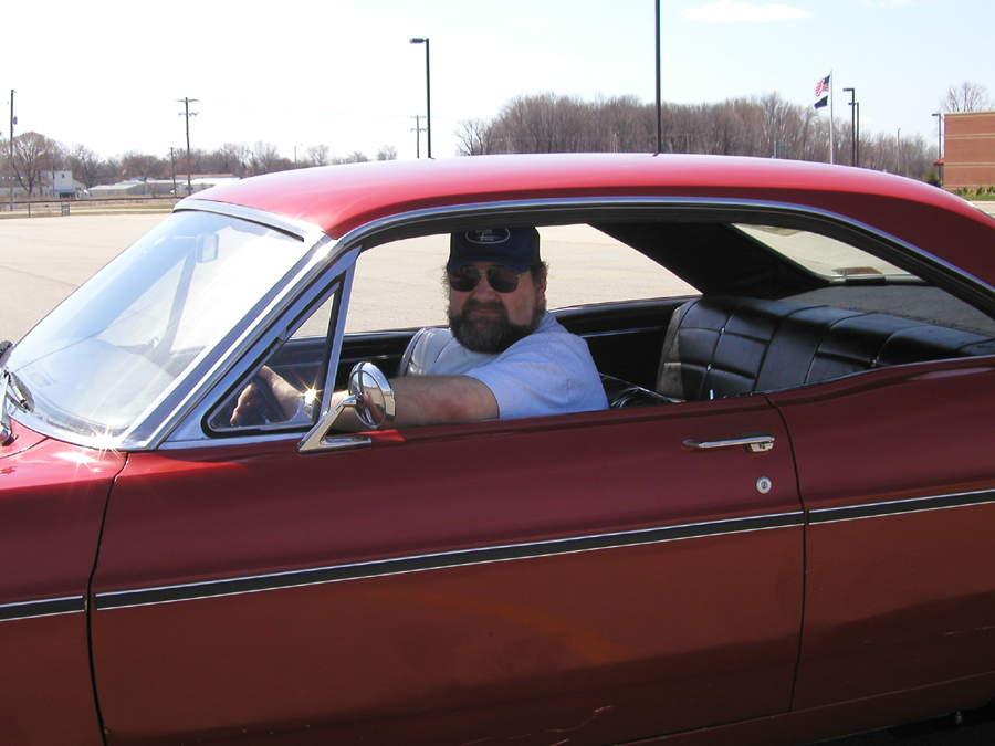 Man in Ford cap and sunglasses sitting in the driver's seat of a red hardtop coupe in a parking lot
