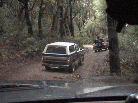 1991 Ford Bronco - Two SUVs driving on a dirt road through a wooded area.