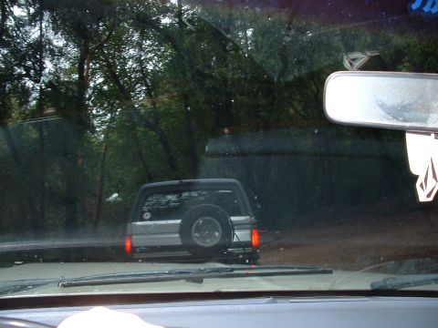 1991 Ford Bronco - A silver Ford Bronco is seen from behind, driving on a dirt road surrounded by trees.