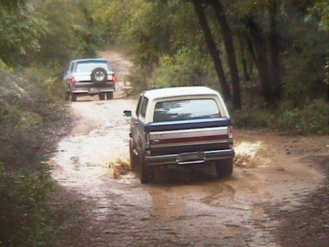 1991 Ford Bronco - Two Ford Broncos navigate a muddy trail through a wooded area.