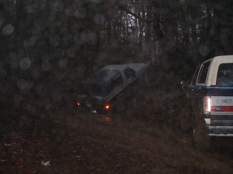 1991 Ford Bronco - Two SUVs navigate a challenging muddy trail in a wooded area, with one vehicle appearing to be stuck.