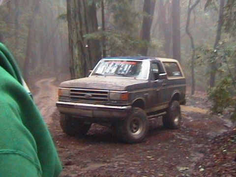 1991 Ford Bronco - Muddy 1991 Ford Bronco navigates a dirt path through a dense, foggy forest.
