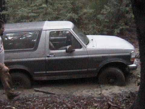 1991 Ford Bronco - A two-tone Ford Bronco is stuck in a muddy, wooded area, with its tires submerged in the mire.