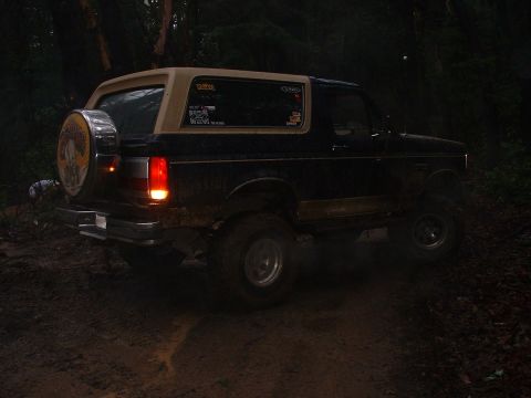 1991 Ford Bronco - A dark 1991 Ford Bronco with a tan top is seen from the rear on a muddy trail in the dark.