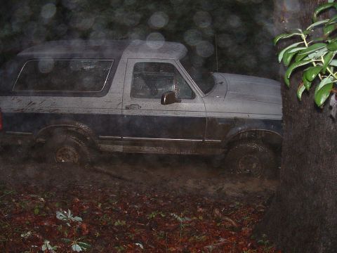 1991 Ford Bronco - Side profile of a two-tone Ford Bronco caked in mud and parked in wet woods at night, shot past a tree trunk with rain drops flaring in the…