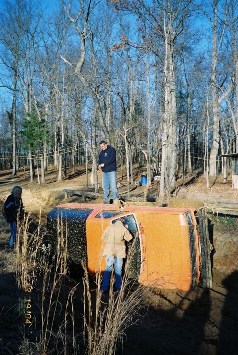 1984 Ford Bronco - An orange SUV has rolled onto its side in a muddy area, with several people observing the scene.