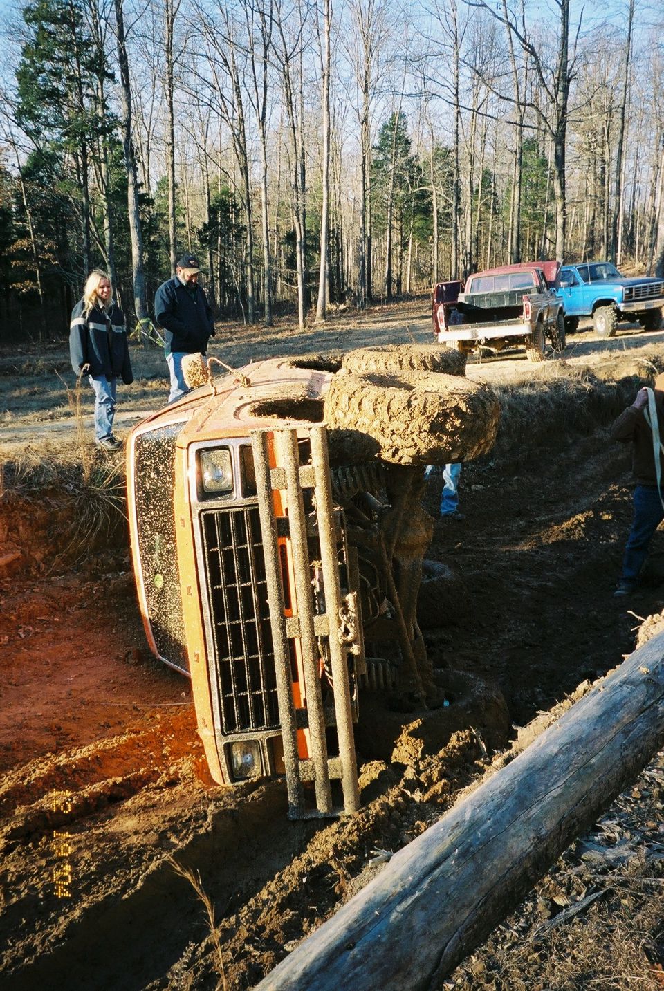 Orange SUV flipped on its side in a muddy ditch, with two people watching and two other trucks in the background
