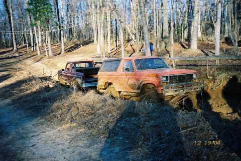 1984 Ford Bronco - Two trucks are stuck in a muddy area with trees in the background. The Bronco is orange and the pickup is maroon.