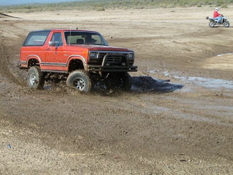 1993 Ford Bronco - Orange Ford Bronco navigates a muddy terrain, with an ATV visible in the distance.