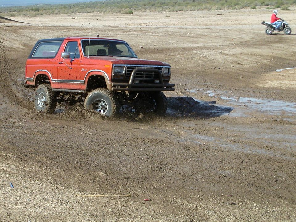 Orange Ford Bronco with aftermarket bumper driving through mud; ATV in background