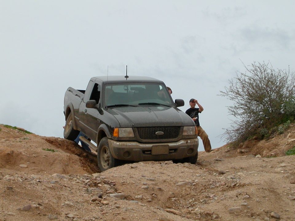 Dark gray Ford Ranger pickup truck navigating a dirt trail with two people visible inside and one standing nearby