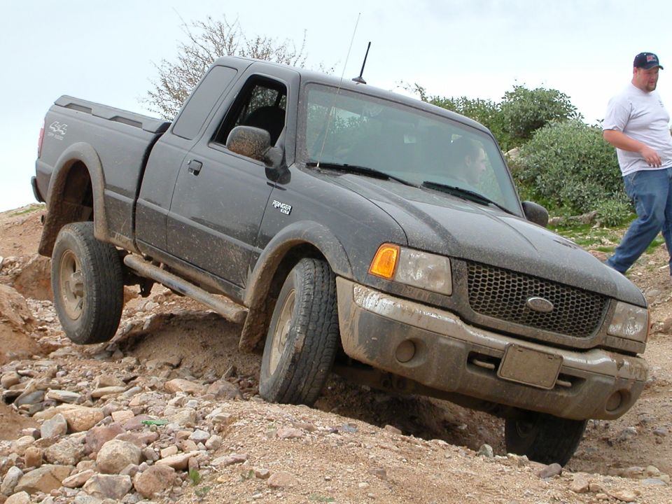Black Ford Ranger pickup truck driving over rocks on a dirt trail. A man walks behind it