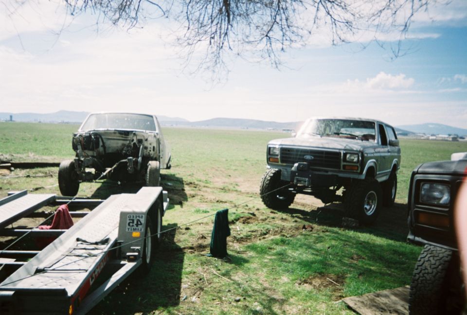 Lifted Ford Bronco on big tires beside a stripped car body on a trailer in a field