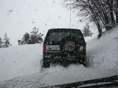 1985 Ford Bronco - A black Ford Bronco navigates a snow-covered road, kicking up snow as it drives.