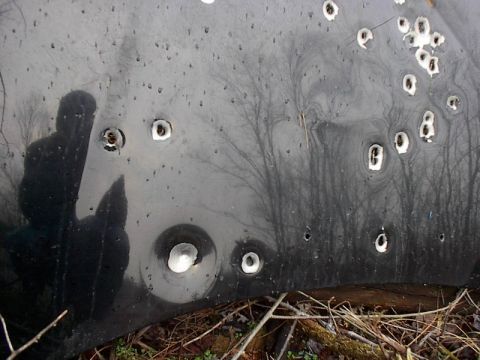 1993 Ford Probe - A black car hood displays extensive damage from bullet impacts, with a reflection of a person visible on its surface.