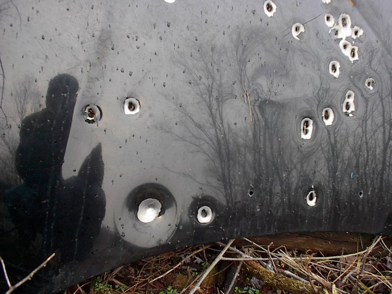 Black car hood with numerous bullet holes, reflection of a person visible
