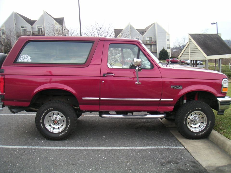 Red Ford Bronco parked on a curb, side view. Lifted suspension and off-road tires are visible