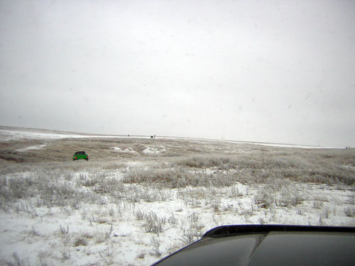 Green SUV parked in a snowy, barren field under an overcast sky. Part of a dark vehicle's dashboard is visible in the foreground