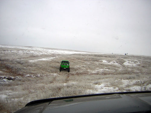 Green off-road vehicle driving across a snowy, grassy field under an overcast sky
