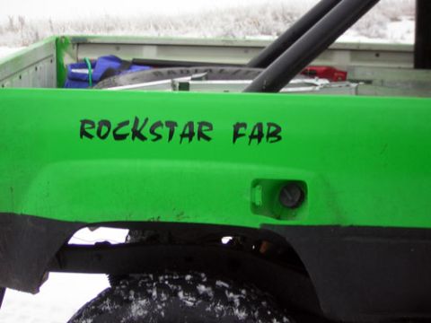 1995 Ford Bronco - The side of a green truck bed displays "ROCKSTAR FAB" lettering and a partial view of a roll bar.
