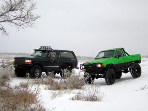 1995 Ford Bronco - A lifted black full-size Bronco with a roof-rack light bar and mud tires sits nose-to-nose in snowy sagebrush with a lifted green compact 4…