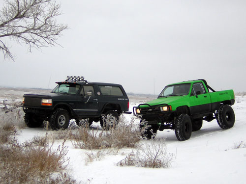 Lifted black Bronco with roof light bar parked in snow beside a lifted green 4x4 pickup
