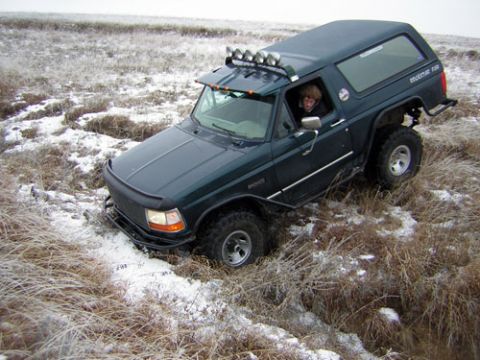 1995 Ford Bronco - Dark green lifted full-size Ford Bronco with a roof-mounted light bar driving through snow-dusted grassland, oversized tires churning the f…