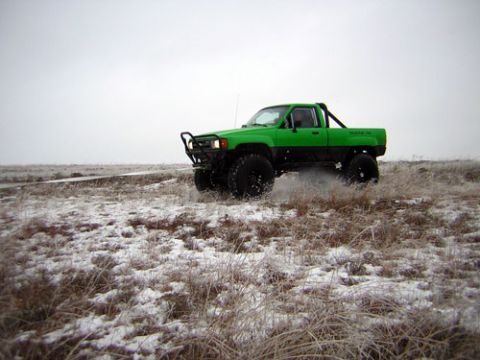 1995 Ford Bronco - A green pickup truck with large off-road tires kicks up snow as it drives through a frosty field.