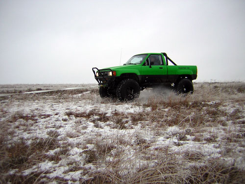Green pickup truck with off-road tires driving through snow-covered field