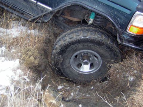 1995 Ford Bronco - Rear corner of a dark green Ford SUV dropped into a snow-dusted ditch, with the passenger rear mud-terrain tire buried in dirt and dead gra…