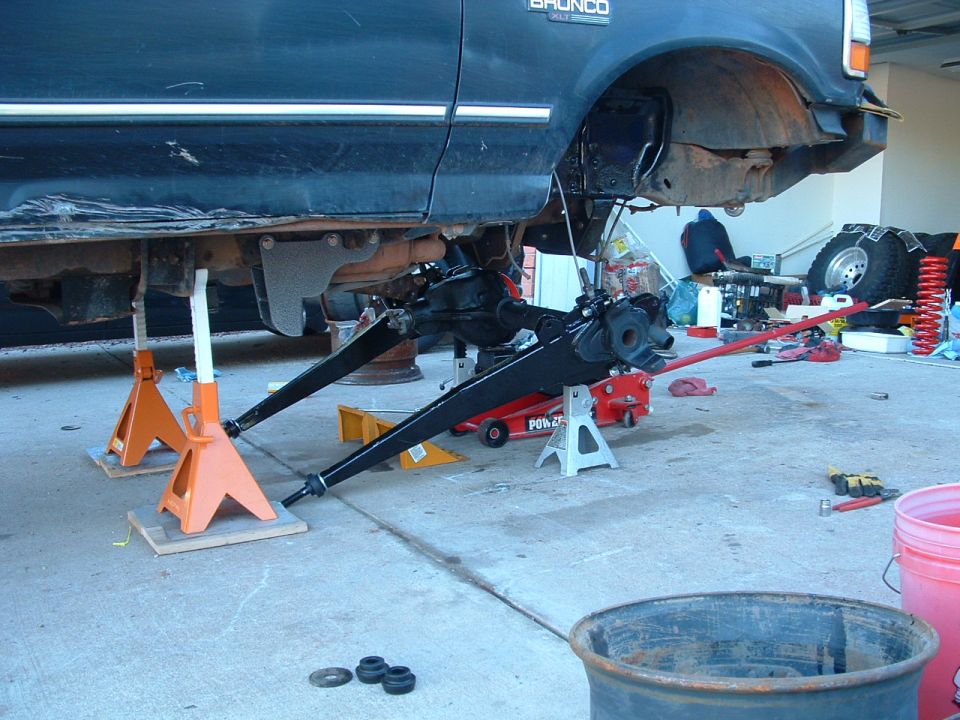 Dark Ford Bronco on jack stands with a black front axle assembly on a floor jack underneath during suspension work