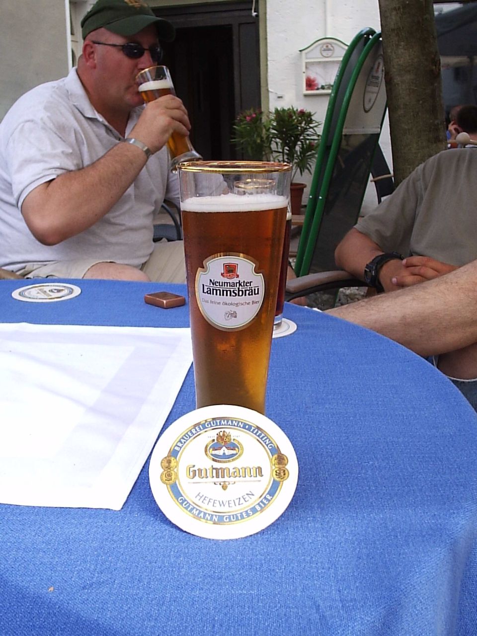 Man in green hat drinks beer next to a glass of Neumarkter Lammsbrau and a Gutmann coaster on a blue tablecloth