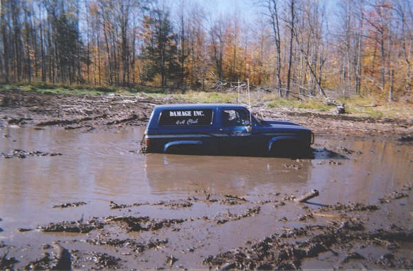 Blue SUV stuck in deep mud with trees in background. Text on door reads 'DAMAGE INC. 4x4 Club'