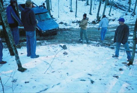 1977 Ford F-150 - Several people stand around a blue sedan that is stuck in a snowy, wooded area.