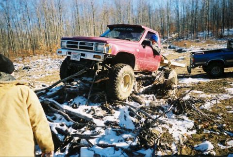 1977 Ford F-150 - A modified red Toyota pickup truck navigates a snowy, wooded area, with another truck visible in the background.