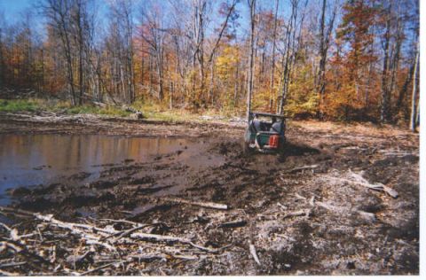 1977 Ford F-150 - A skid steer loader is stuck in a muddy area with a pond and trees visible.