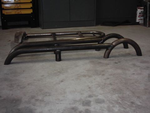 1978 Ford Bronco - A shop-fabricated tubular bumper assembly in bare welded steel sitting on a concrete garage floor, showing bent hoops, cross tubes, a light…