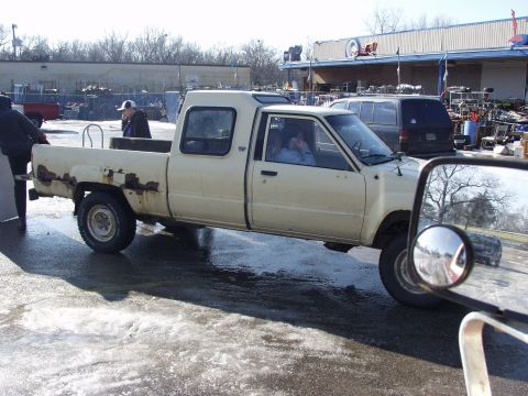 1979 Ford Bronco - A cream-colored pickup truck with a camper shell is parked on a wet surface, with other vehicles and buildings in the background.