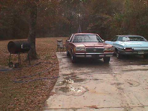 1994 Ford Crown Victoria - A red 1994 Ford Crown Victoria is parked in a driveway next to a blue car, with a barbecue grill visible on the left.