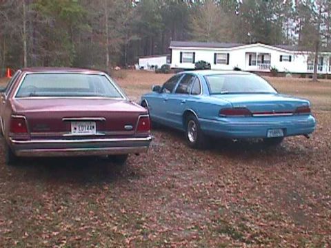1994 Ford Crown Victoria - Two sedans are parked on a gravel lot in front of a mobile home, with trees in the background.