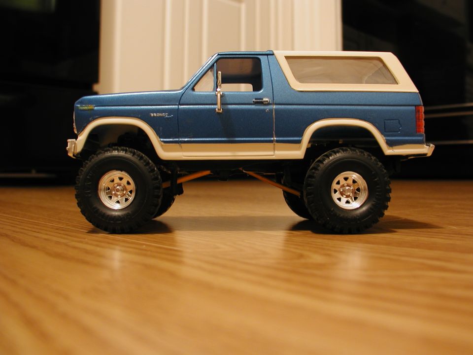 Blue and white Ford Bronco scale model in profile on a wood floor with lifted off-road tires