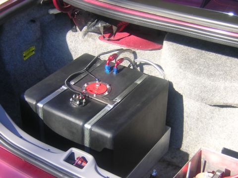 1992 Ford Crown Victoria - A black plastic fuel cell strapped down with polished aluminum bands in the gray carpeted trunk of a maroon car, topped by a red anodized f…