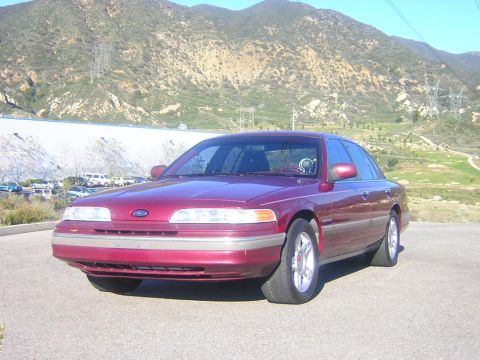 1992 Ford Crown Victoria - A maroon/burgundy 1992 Ford Crown Victoria four-door sedan photographed from the front three-quarter angle, parked on pavement with mountai…