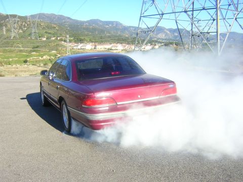 1992 Ford Crown Victoria - A dark red/maroon Ford Crown Victoria sedan viewed from the rear three-quarter angle doing a burnout, with thick white tire smoke billowing…