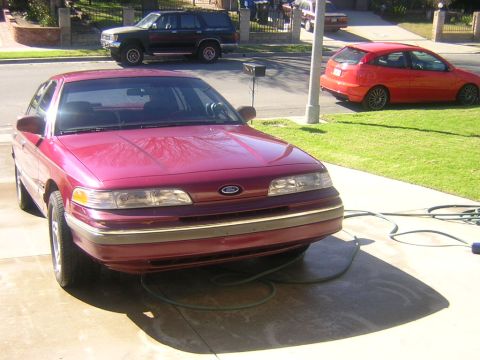 1992 Ford Crown Victoria - A dark red/burgundy Ford full-size sedan photographed from the front three-quarter angle in a driveway, with a green garden hose coiled on…