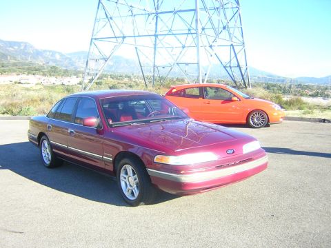 1992 Ford Crown Victoria - A maroon/burgundy 1990s Ford Crown Victoria sedan shown from a front three-quarter angle, parked on asphalt with a red interior visible and…