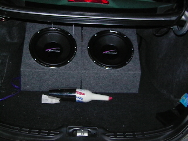 Two car subwoofers mounted in a carpeted enclosure in a trunk, with a JBL spray bottle resting on the floor