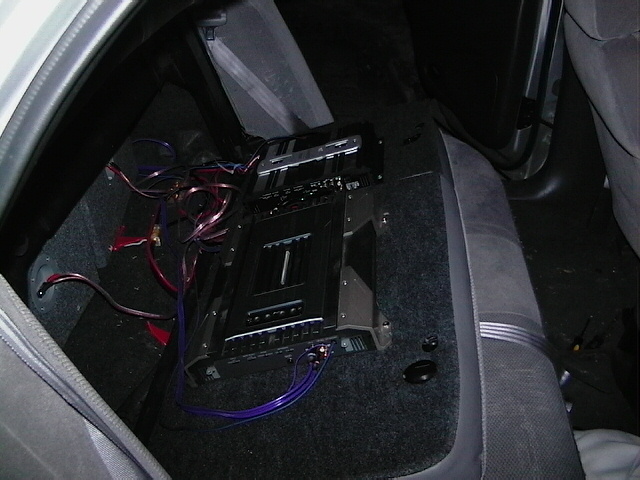 Car interior showing an audio amplifier installed in the rear seat area with visible wiring